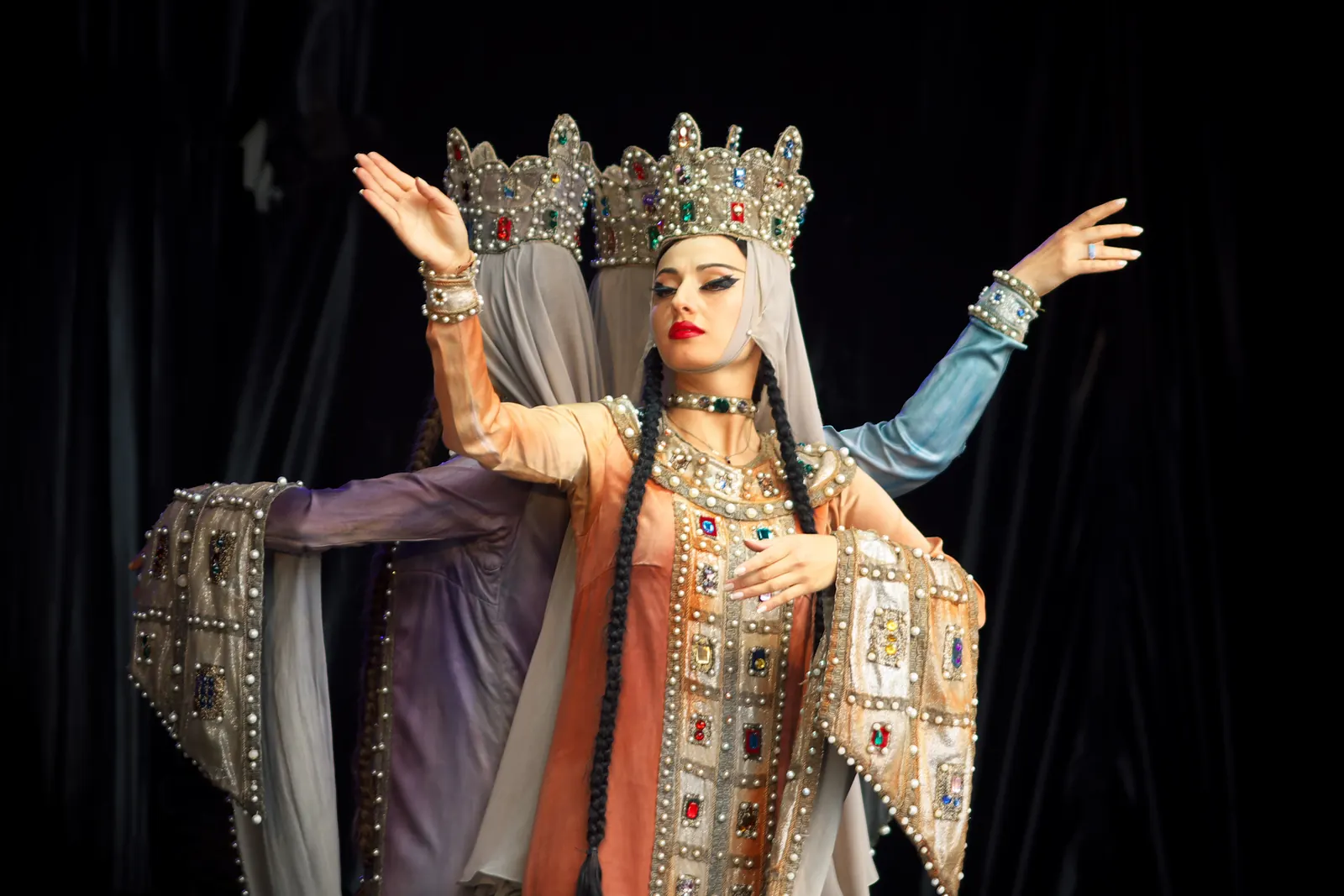 Three Georgian women dancers in jewelled crowns and long beaded sleeves, one in front with her arms raised, on a dark stage