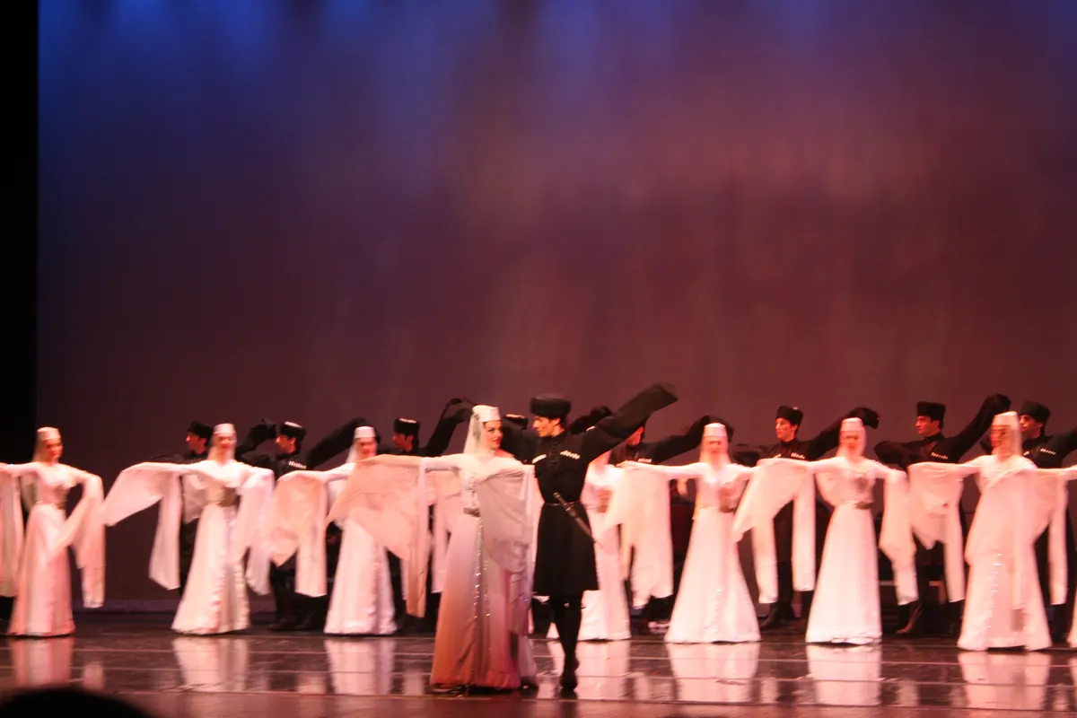 A line of Georgian dancers, men in dark coats and women in white with long veils and wide sleeves, on a stage