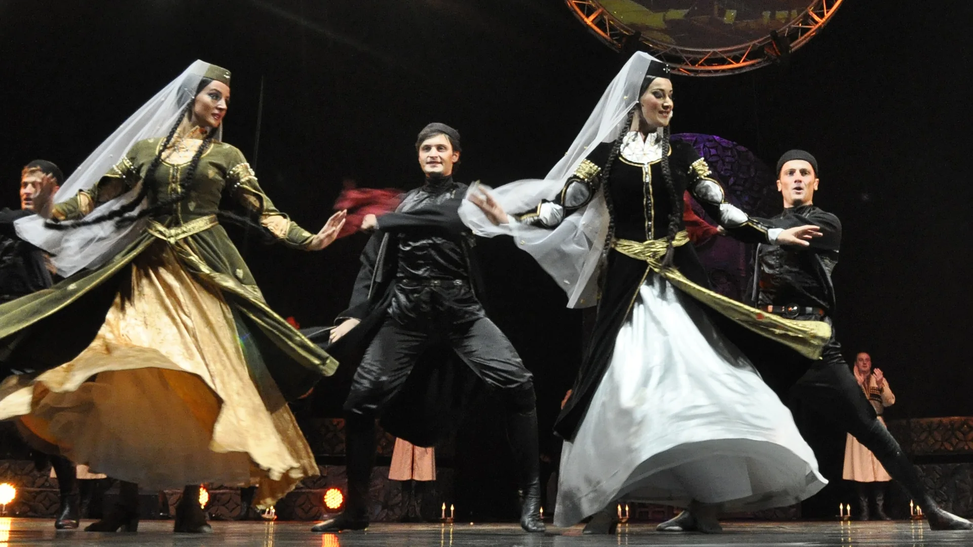 Four Georgian folk dancers in black, gold and white costumes with long veils mid-turn on a dark stage, candles glowing along its edge